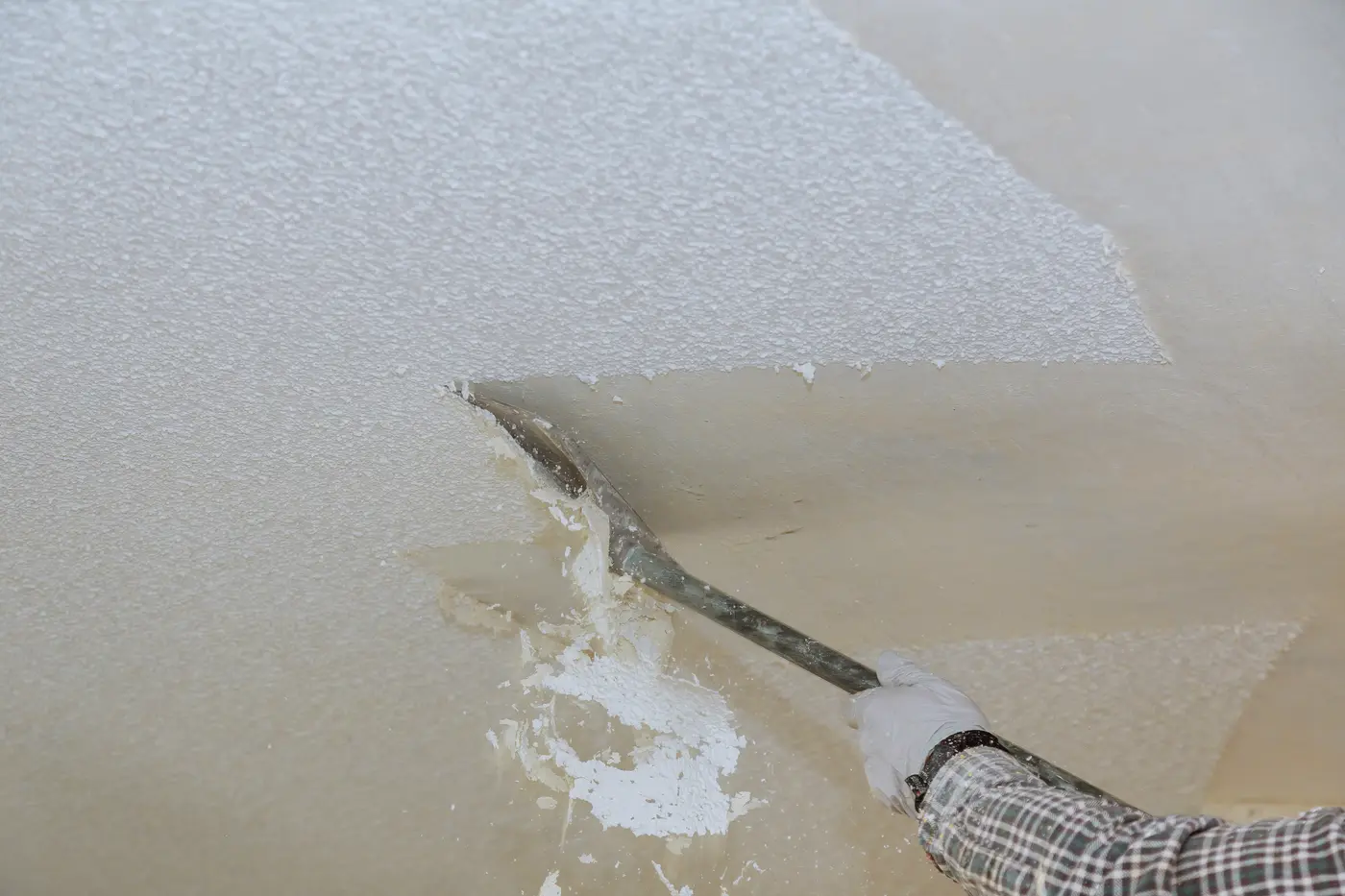 Gloved hand using a long-handled scraper to strip popcorn texture off a ceiling